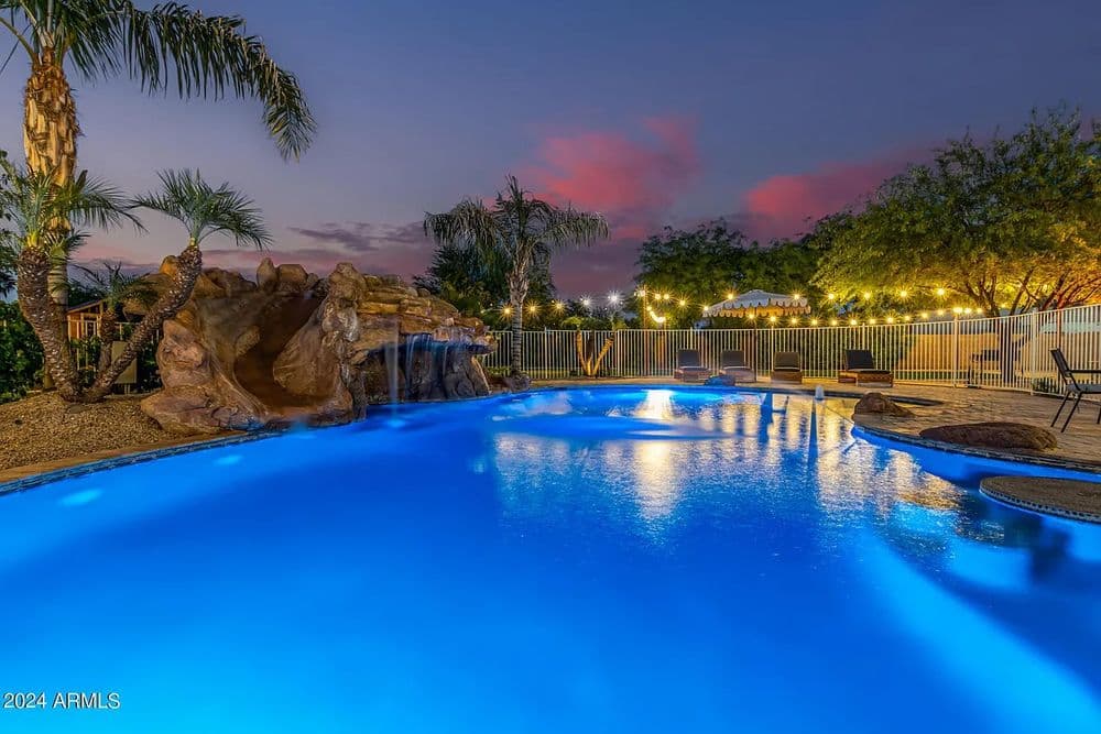 Vibrant illuminated swimming pool with waterfall and palm trees at twilight.