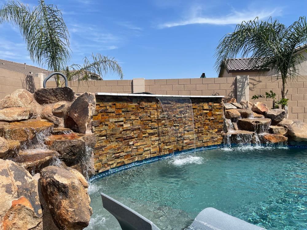 Modern backyard swimming pool with a stone waterfall feature and palm trees under a blue sky.