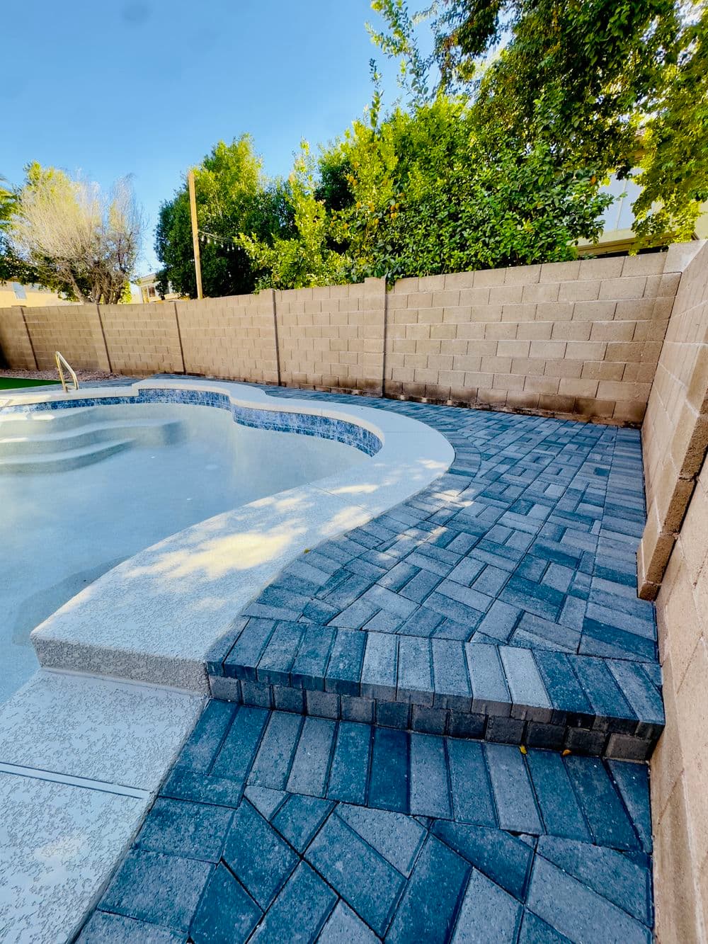 Backyard pool area with patterned paver stones and landscaped greenery in the background.