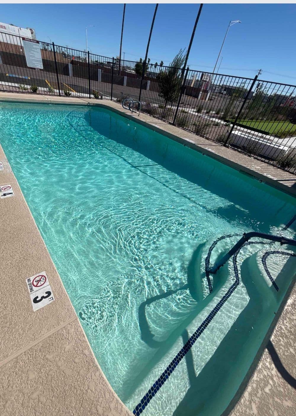Clear swimming pool with stairs, surrounded by a fence and landscaped area under a blue sky.