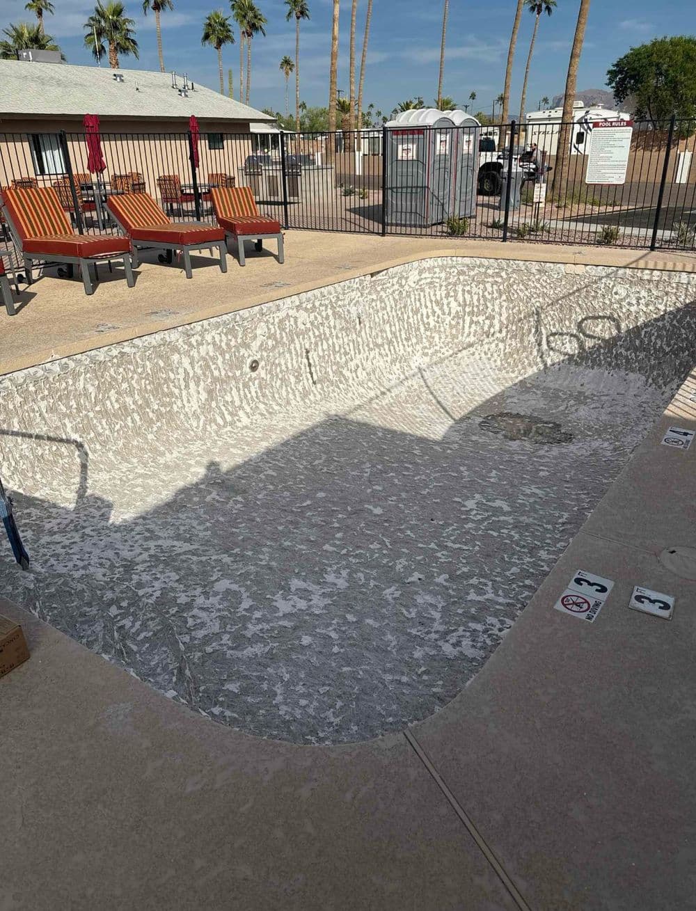 Empty swimming pool with textured concrete surface, surrounded by palm trees and lounge chairs.