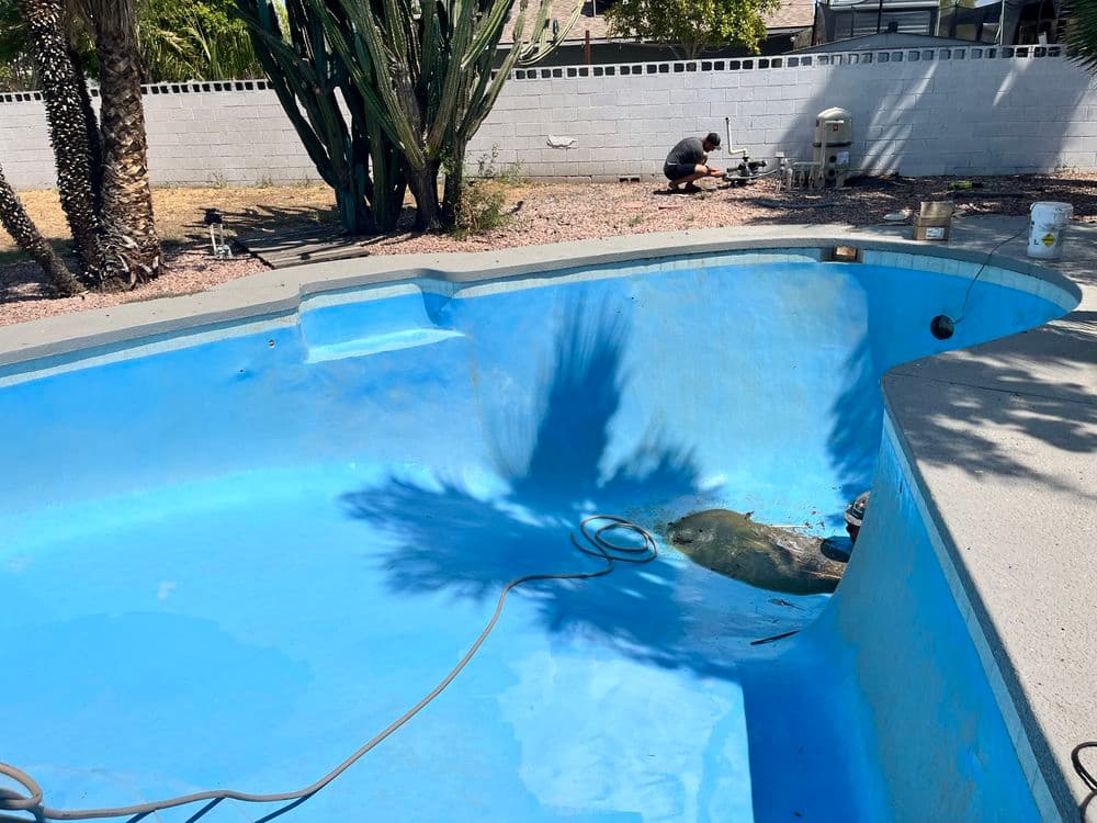 Empty swimming pool being repaired, with shadows cast from palm trees and a worker in the background.