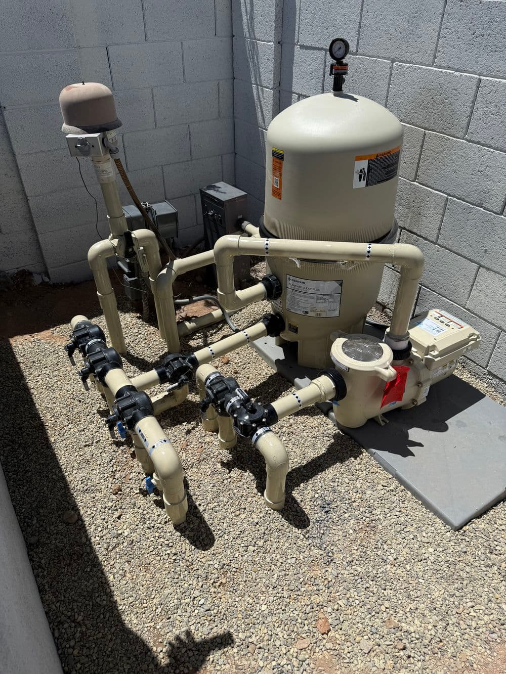Swimming pool filtration system with cream-colored pump, pipes, and valves in a gravel area.
